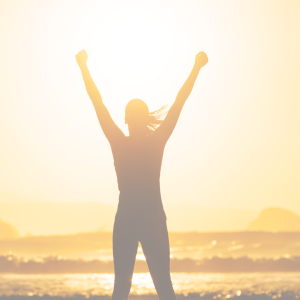 woman standing at beah and sunset raising her hands above head denoting success