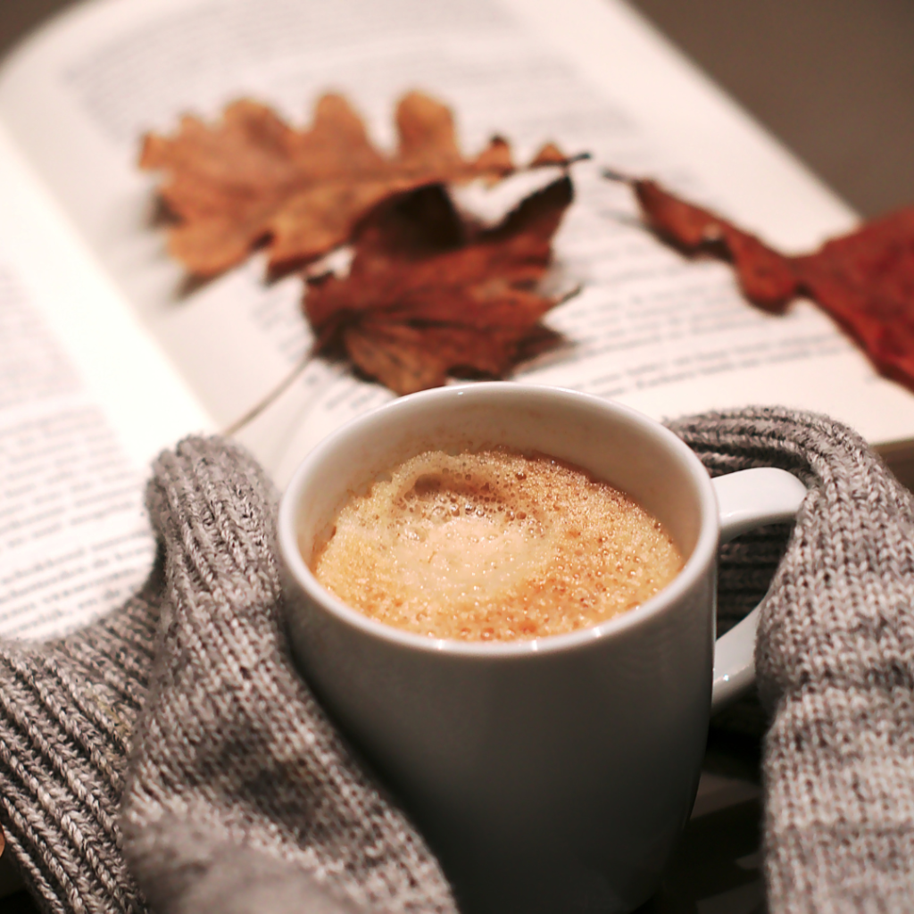 a pair of hands holding onto a hot mug with a book open in the background with a few autumnal leaves on the page